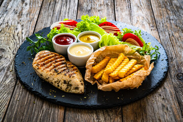 Grilled chicken breast, French fries and fresh vegetables on wooden table
