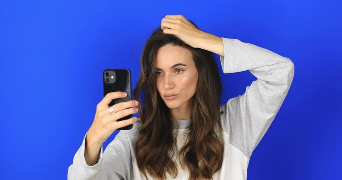 The girl stares at the phone and fixes her hair. Female uses phone like mirror. Young girl make photo from hands with phone on blue background, look at camera and smiling.