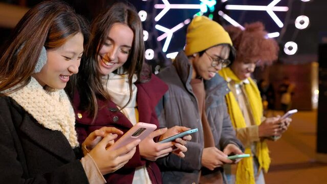 Young happy friends standing in a row having fun together using cell phones on city street at night on winter season. Teenage people watching social media content on mobile app. Front view footage.