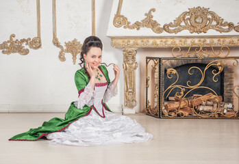 Young beautiful smiling woman in green rococo style medieval dress sitting near fireplace