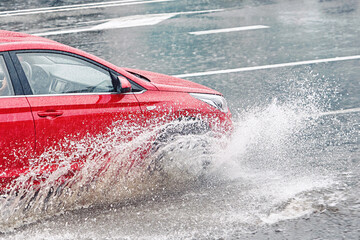 Red car driving through puddle during heavy rain, mooving fast on wet road. Red car driving on flooded asphalt road. Dangerous driving, risk of aquaplaning.
