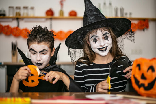 Cute brother and sister are sitting in the kitchen at the table, made up in witch and vampire costumes, making crafts, cutting out paper, preparing for Halloween. Children making crafts for party.