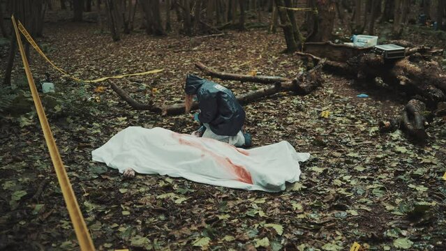 Coroner ispecting a dead body and taking dna samples from a Crime Scene in the woods. Cadaver covered in bloody white sheet
