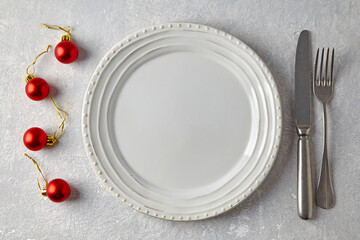 Empty white plate, red Christmas balls and cutlery on a gray stone table. New Year's menu