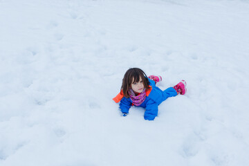 Fototapeta na wymiar child in bright blue winter coveralls lying down on the snow and watching at camera
