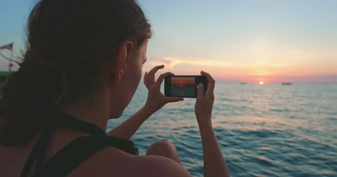 Tourist woman making photo of colorful ocean sunset while having boat tour to Phi Phi Islands, Thailand. Close up caucasian girl back view holding mobile phone take landscape panorama. Slow motion 4K