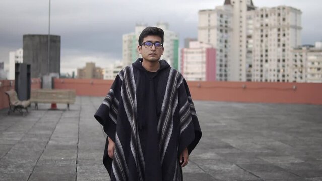 Joven con gafas vistiendo poncho negro boliviano (vestimenta tradicional/tipica)andino, folcl&oacute;rico, ciudad de fondo con edificios al atardecer