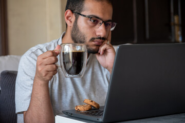 bearded male drinks coffee while working on his laptop from his home and eating cookies