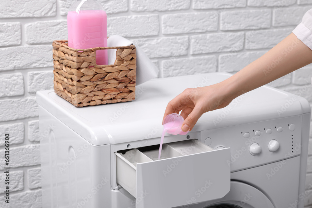 Wall mural Woman pouring fabric softener from cap into washing machine near white brick wall, closeup