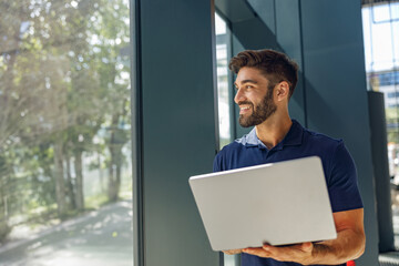 Smiling male sales manager working on laptop standing in office during working day and looks away