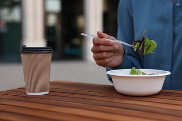 Businesswoman with plastic bowl of salad and paper cup of coffee having lunch at wooden table outdoors, closeup