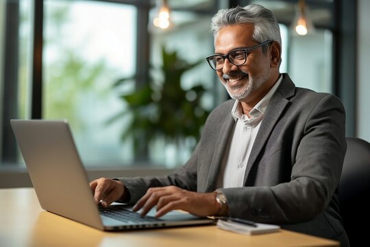 A smiling, busy mid-aged professional businessman works on his laptop at his office desk. An older, mature Indian businessman, a happy male entrepreneur worker, types on his computer
