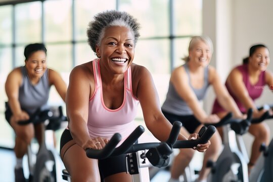 Group of women of different ages and races during cycling workout. Group fitness classes on exercise bikes. Workouts for any age. Be healthy in any age. Photo against a bright, gym studio background.
