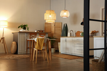 Interior of modern kitchen with glowing lamps, dining table and white counters at evening - obrazy, fototapety, plakaty