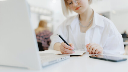 Young beautiful joyful blonde influencer woman blogger writes content plan in notepad in cafe with coffee, white light