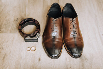 Men's brown leather stylish groom's shoes, black belt, gold rings lie on the floor. Close-up wedding photo, top view.