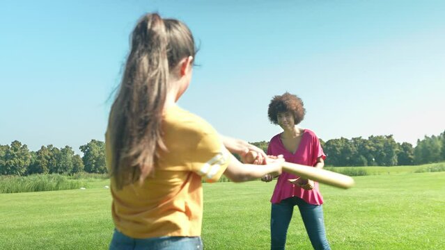 Positive attractive African American mother with curly hair and lovely multiracial teenage girl enjoying leisure and sport activities together, practicing baseball game on green field in public park.