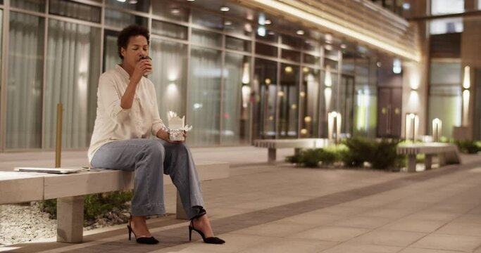 Ethnic businesswoman drinking coffee outside office building in evening