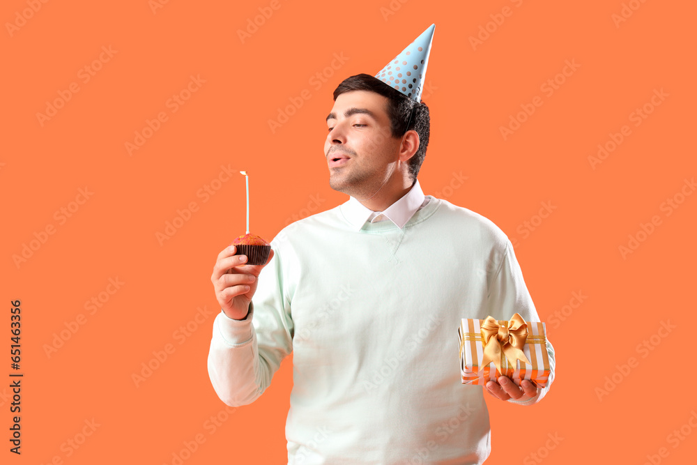 Sticker Happy young man with birthday muffin and gift making wish on red background