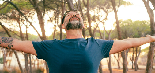 Front view of happy overjoyed mature man outstretching arms and enjoying outdoor leisure activity with park trees in background. People and happiness. Joyful male opening arms hugging life. Joyful