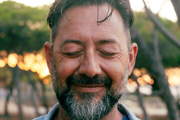 Close up portrait of mature man with beard and closed eyes smiling on camera. Wellbeing and mental...