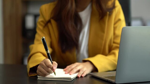 Business woman using laptop to work and writing on notebook with pen in office with mobile next to laptop.
