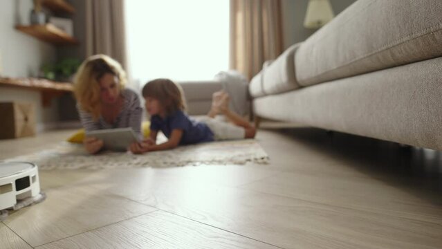 Vacuuming robot cleaning at home. Wireless robot vacuuming in the living room. Blurred family mom and son laying on floor on carpet while vacuum cleaner working. Domestic appliance and technology.