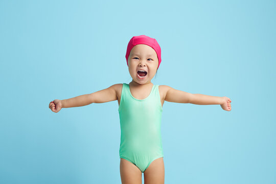 Little female child swimmer cheerfully spreads her arms to the sides, dressed in a turquoise swimsuit and a pink cap standing on a blue isolated - Powered by Adobe