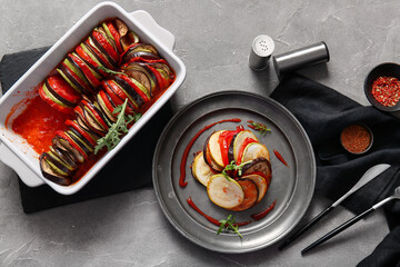 Baking dish and plate of tasty Ratatouille on grey background