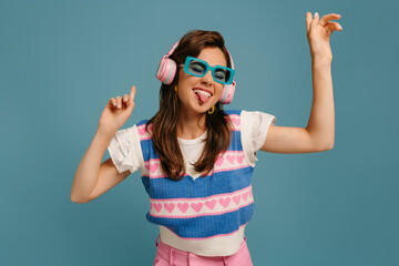 Playful young woman in headphones and trendy glasses enjoying music while dancing on blue background