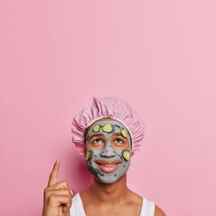 Vertical shot of pleased African man undergoes beauty procedures at home points index finger above shows blank space for your promotion applies nourishing facial clay mask with cucumber slices