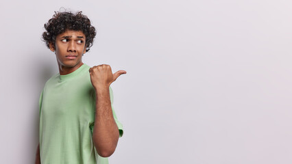 Horizontal shot of curly haired young Hindu man turns back and points thumb aside on copy space shows mock up space for your promotional content isolated over white background. Look at this.