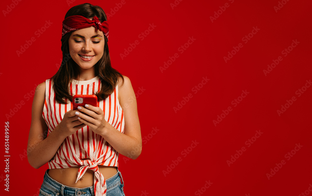 Fototapete stylish young hipster woman texting message on smart phone and smiling while standing on red backgro