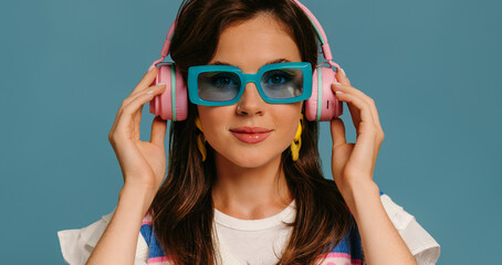 Portrait of beautiful young woman in headphones and trendy glasses enjoying music on blue background