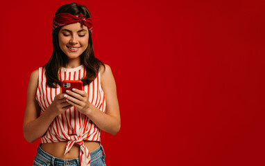 Stylish young hipster woman texting message on smart phone and smiling while standing on red background