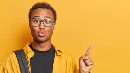 Photo of handsome curly haired African man indicates index finger on copy space shows something at upper right corner dressed in casual shirt isolated over yellow background. Follow this way