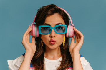 Portrait of fashionable young woman in headphones and trendy glasses enjoying music on blue...