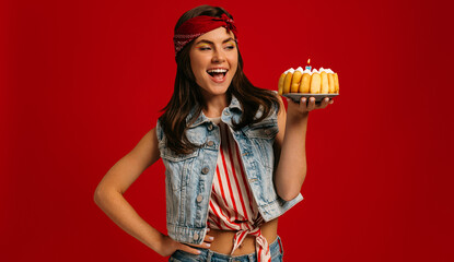 Happy young woman holding birthday cake and looking at it with smile on red background
