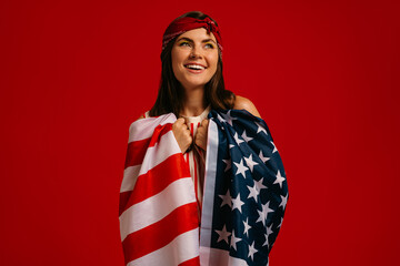 Fototapeta premium Attractive young hipster woman carrying American flag on shoulders and smiling on red background