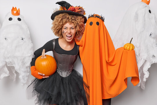 Having Halloween party concept. Indoor shot of young beautiful terrifying African american model wearing dress and hat of witch holding pumpkin standing among spooky ghosts looking straight at camera