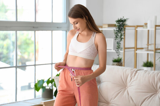 Beautiful young woman measuring her waist in living room. Weight loss concept