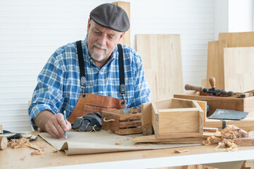 Caucasian senior old white bearded man carpenter in apron, hat and gloves working in workshop, use pencil sketching a design on paper, handmade wood toy, tools and handcrafts is on table in white room