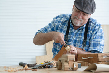 Caucasian senior old white bearded man carpenter in apron and hat using chisel working in workshop,...