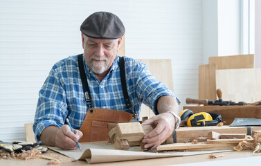Caucasian senior old white bearded man carpenter in apron and hat working in workshop, sketching a...