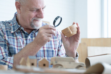Caucasian senior old white bearded man carpenter working in workshop, holding magnifying glass look...