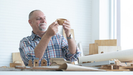 Caucasian senior old white bearded man carpenter working in workshop, holding, looking at handmade...
