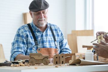 Selective focus on car and house wooden toy on table. Caucasian senior old white bearded man carpenter in apron and hat working in workshop, sketching on paper in white room