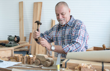Caucasian senior old white bearded man carpenter using hammer and chisel working in workshop, tools machine small wooden toy model or handcrafts is on table in white room. Professional carpentry
