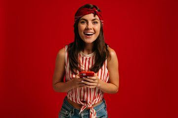 Joyful young hipster woman holding smart phone and smiling while standing on red background