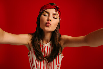 Beautiful young hipster woman making selfie while keeping arms outstretched on red background